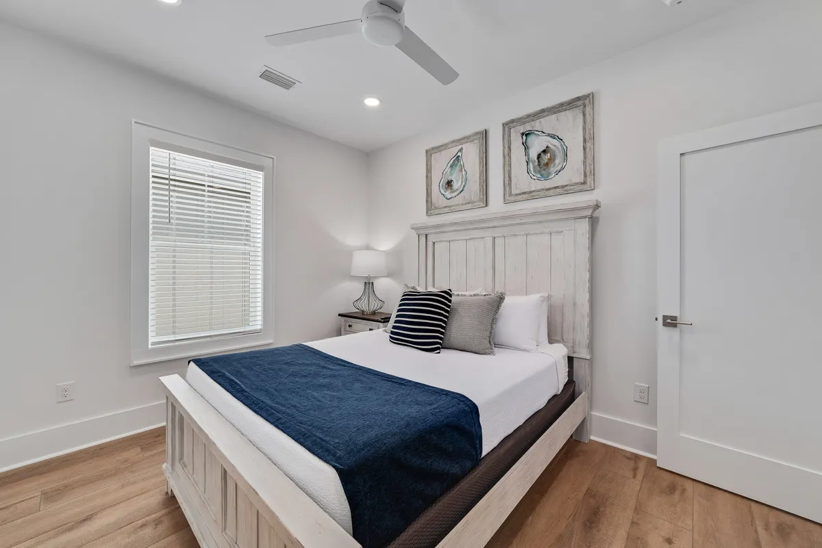 A bright, serene bedroom with crisp white and navy bedding, a wooden bedside lamp, and a window with blinds letting in natural light. The room feels meticulously ordered and fresh, with framed oyster shell art providing a subtle coastal touch. Surfaces appear spotless and linens immaculate, echoing hotel-level standards.