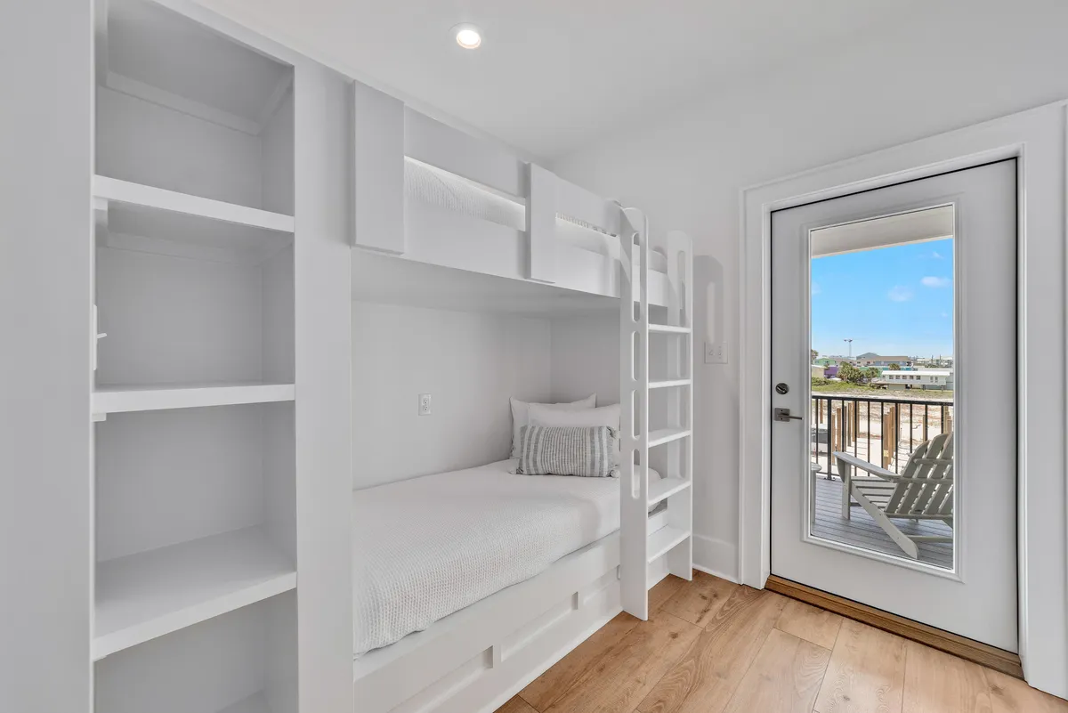A cheerful bunk nook with two bunk beds, a large window streaming in natural light, minimalist shelving, and wood flooring. The playful setup is perfect for children or cousins to share stories and laughter.