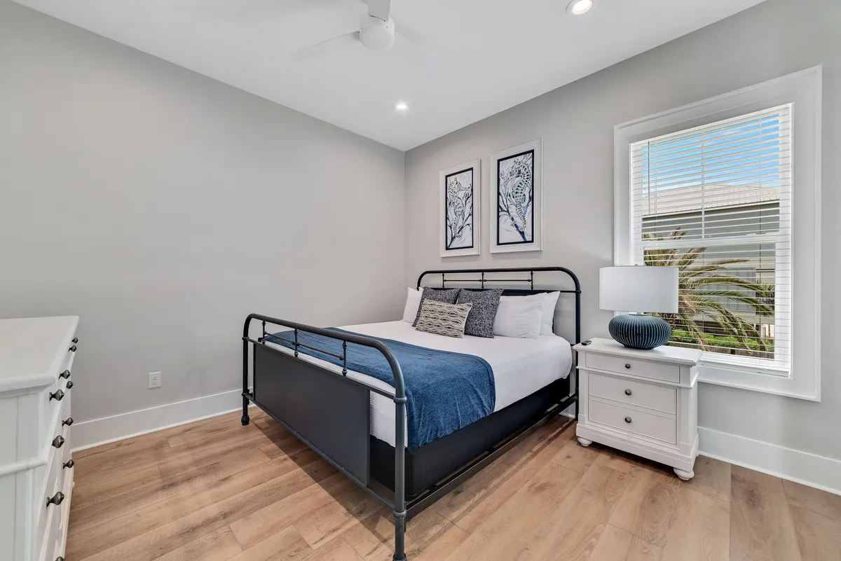 A serene bedroom featuring a bed with a blue throw blanket, framed artwork on the wall, and a nightstand with a lamp. Soft natural light enters through the window, highlighting the tranquil atmosphere and inviting bedding.