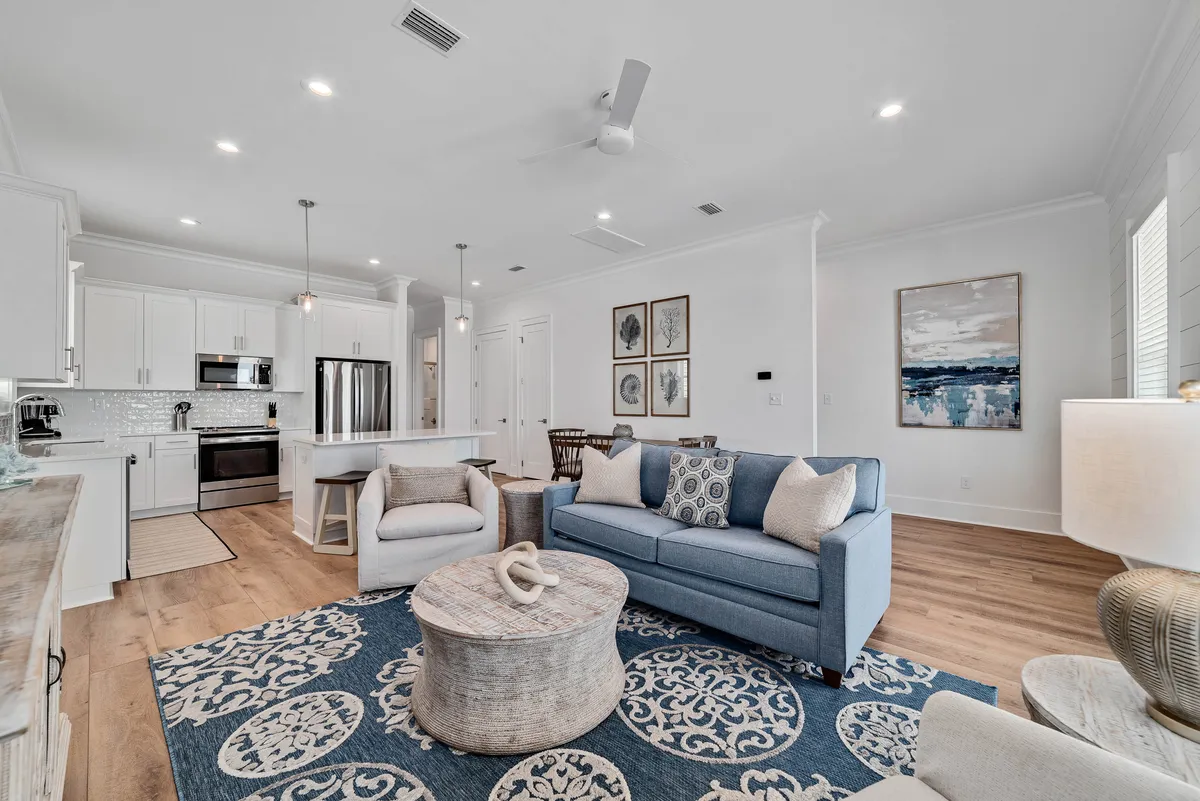 The image showcases a bright and airy open-concept living area featuring a stylish blue patterned rug, a comfortable light blue sofa adorned with decorative pillows, a round wooden coffee table, and a modern kitchen with white cabinets and a spacious island. The space feels inviting and perfect for family gatherings, relaxation, and shared meals.