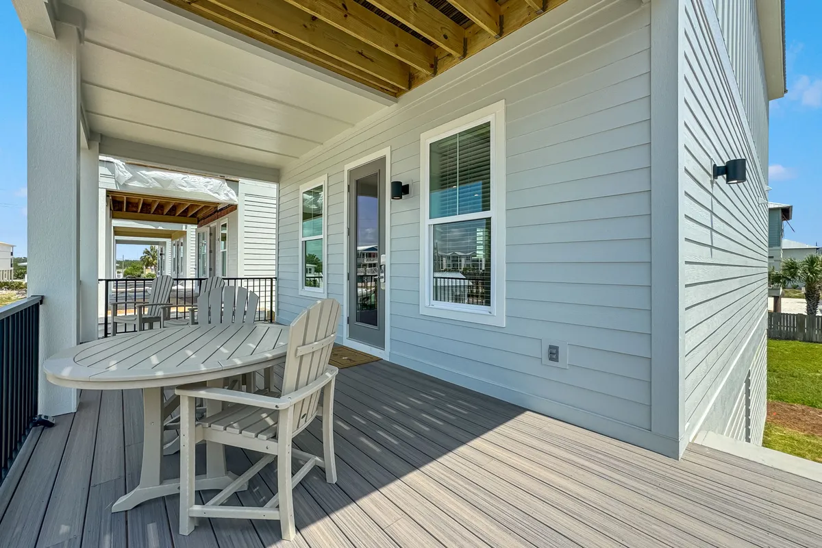 A covered balcony with a white wooden table and chairs set against light blue exterior siding, offering views of neighboring houses and lush greenery. This inviting space is ideal for outdoor dining, morning breakfasts, or evening conversations, capturing the essence of alfresco living.