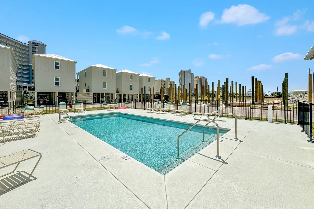 A vibrant outdoor scene featuring the community swimming pool with clear blue water, surrounded by lounge chairs and modern elevated beach houses in the background. The area is designed for both relaxation and socializing, offering a lively yet tranquil atmosphere for guests and families.