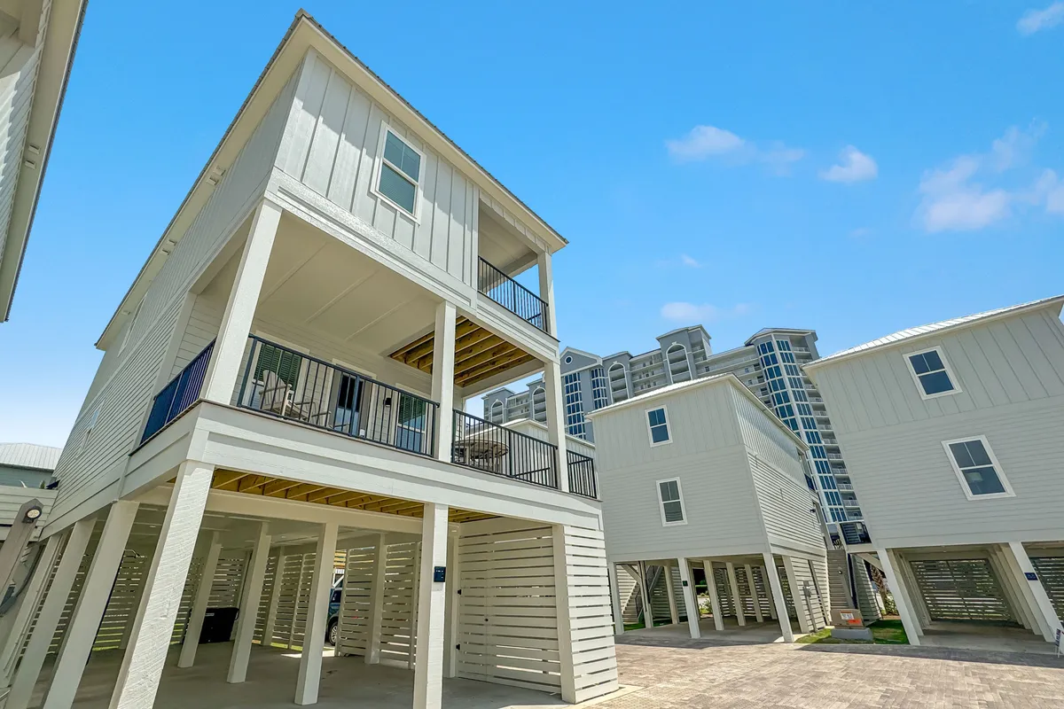 This image captures elevated modern beach houses on stilts with inviting balconies, open parking areas, and a clear blue sky overhead. The scene radiates a relaxed coastal vibe, hinting at the close proximity to the beach and the welcoming neighborhood atmosphere.
