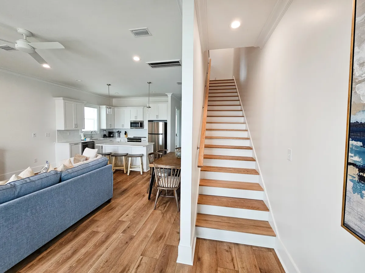 An open-concept interior with a white kitchen cabinetry and island, adjacent to a wooden dining table and chairs. The blue couch and a wooden staircase leading to the upper floor are visible in the background, illustrating the home’s open layout and integrated living, dining, and kitchen spaces.