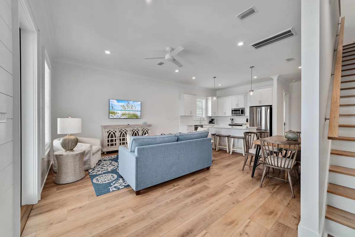 A wide view of a light blue sofa anchoring the living area, patterned rug, and a modern white kitchen with stainless steel appliances. The wooden dining table and chairs are positioned near a staircase, emphasizing the open-concept flow between living, dining, and kitchen spaces.