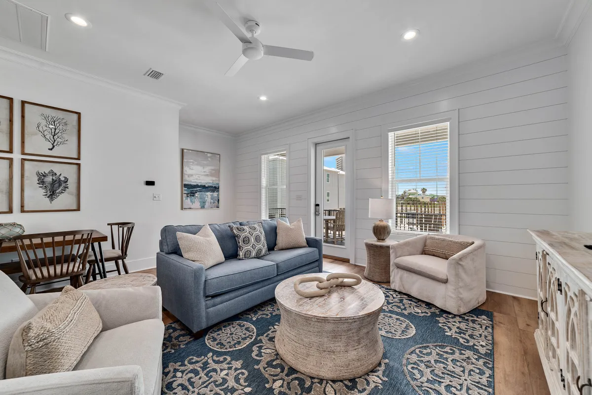 A cozy living room featuring a blue upholstered sofa with patterned pillows, a round wooden coffee table with a decorative centerpiece, and two beige armchairs. The area is anchored by a large blue and beige patterned rug, and there's a glass door offering views and access to an outdoor balcony. The adjacent dining area is visible, providing a sense of open-concept flow.