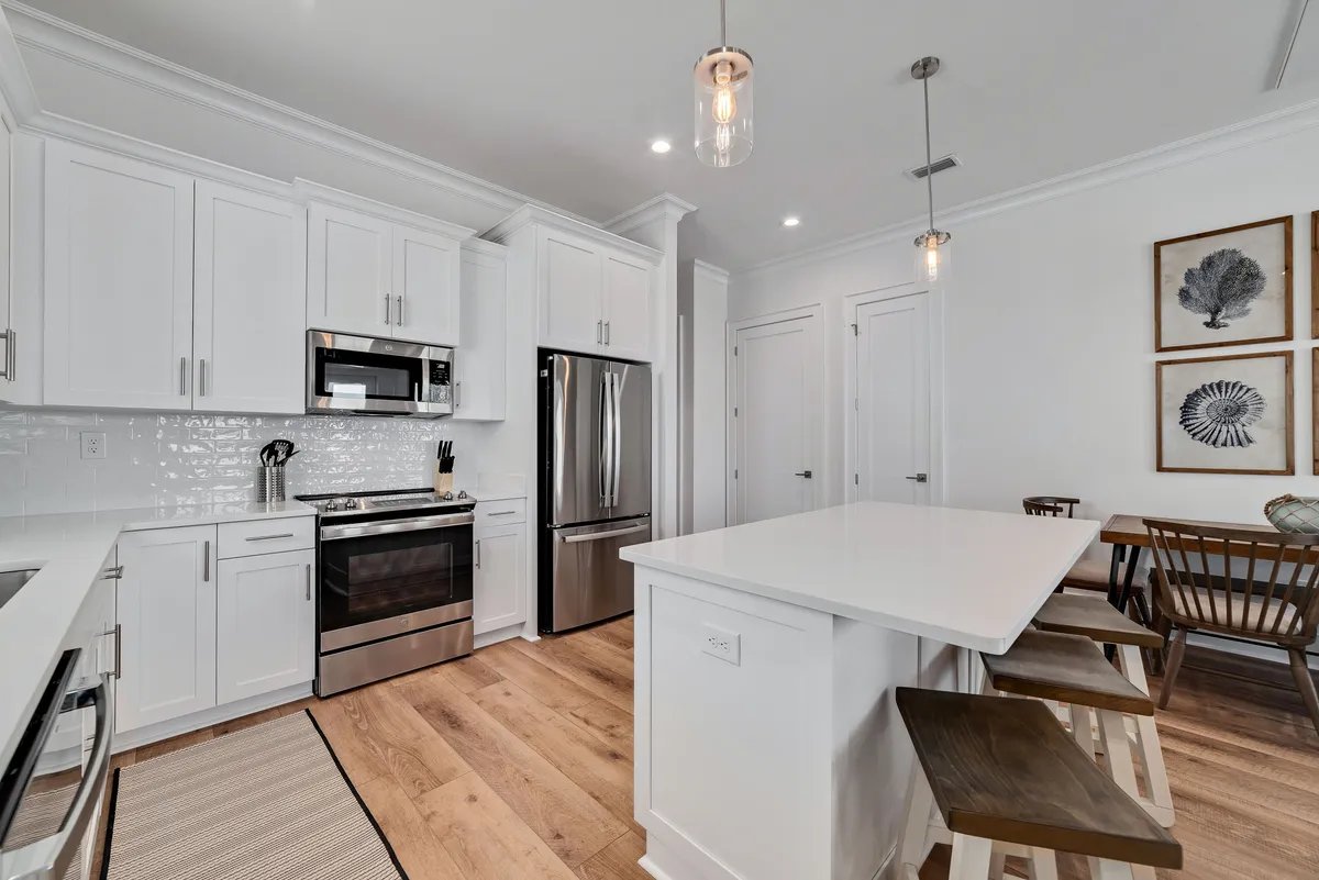 A cozy, functional dining and kitchen area featuring a wooden 4-seater dining table and a kitchen island with stools. Stainless steel appliances and white cabinetry with a subway tile backsplash create a fresh, modern look. The space is filled with natural light, encouraging family meals and togetherness.
