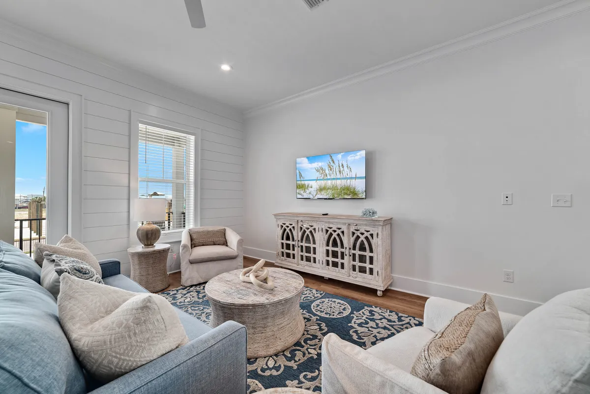 A cozy living room featuring a light blue sofa with neutral cushions, white armchairs, a rustic round coffee table, and a decorative media cabinet with a TV. Natural light streams in, suggesting a connection to the outdoors and a calming, beach-inspired atmosphere ideal for casual conversations or group relaxation.