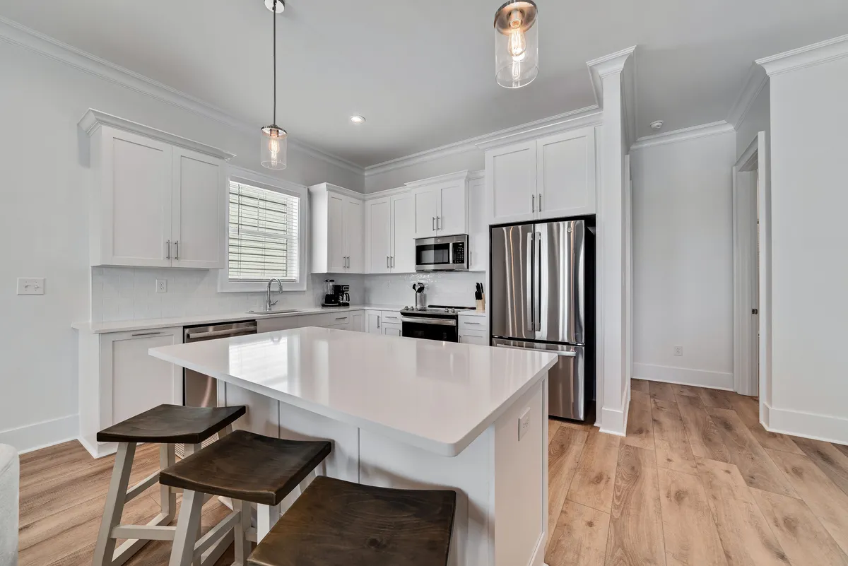 A bright, stylish kitchen featuring a large white island bar at the center, flanked by wooden stools. Modern stainless steel appliances line the background, while pendant lighting above the island adds a welcoming glow. The open layout invites interaction, with plenty of counter space for meal prep and casual dining.