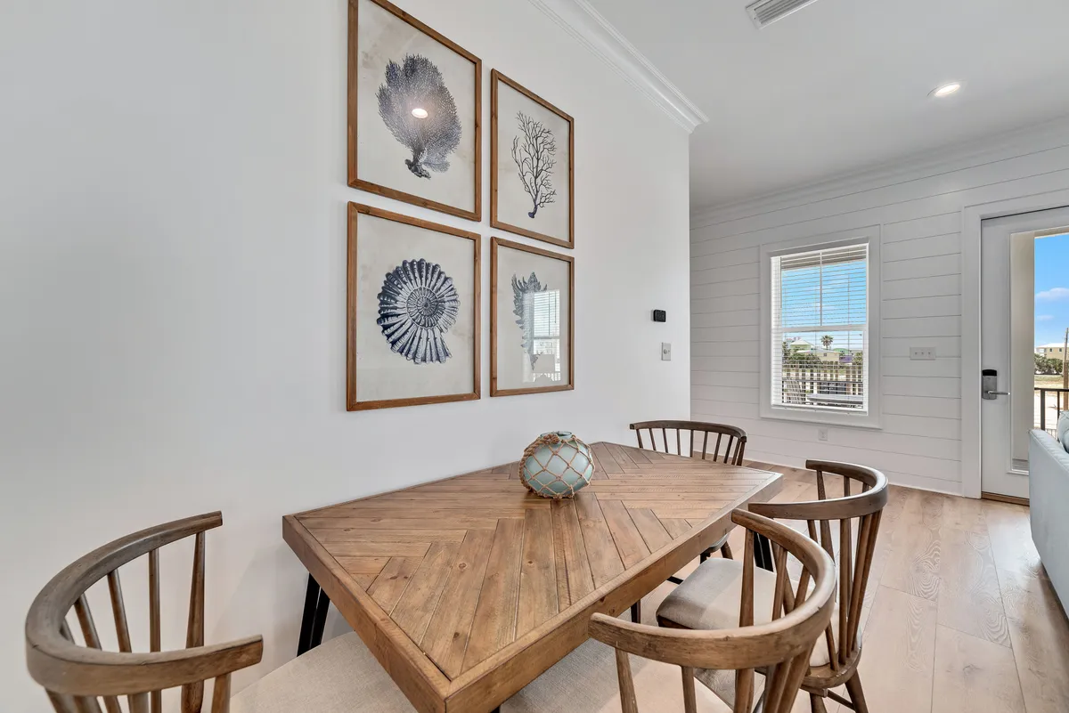 A bright and inviting dining area with a wooden dining table featuring a geometric pattern, surrounded by four rustic wooden chairs. Nautical-themed artwork and a window with a glass door allow natural light to pour in, highlighting the space's coastal décor and relaxed sophistication.