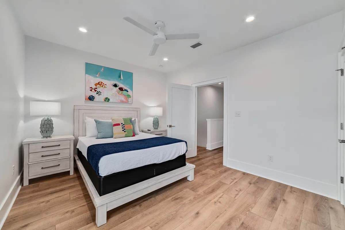 A plush queen size bed dressed in high-quality linens, flanked by matching bedside tables and lamps. Above the bed, beach-themed artwork adds a coastal touch, and the tranquil mood is complemented by soft natural lighting.