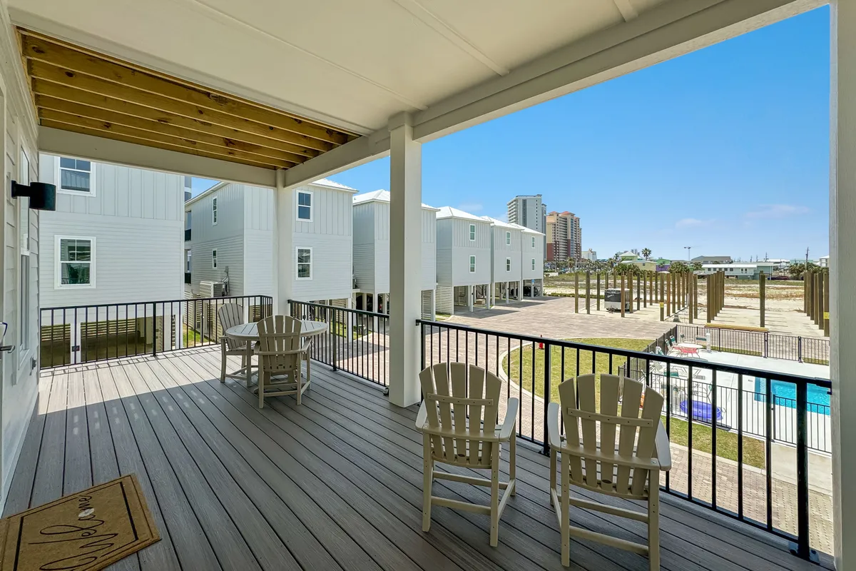 This image shows a covered balcony deck furnished with Adirondack chairs and a small table, overlooking the community pool below. The distant multi-story buildings and open sky create a sense of openness and connection to the outdoors. The balcony setting is perfect for morning coffee or evening drinks, with easy sightlines to the pool and communal spaces.