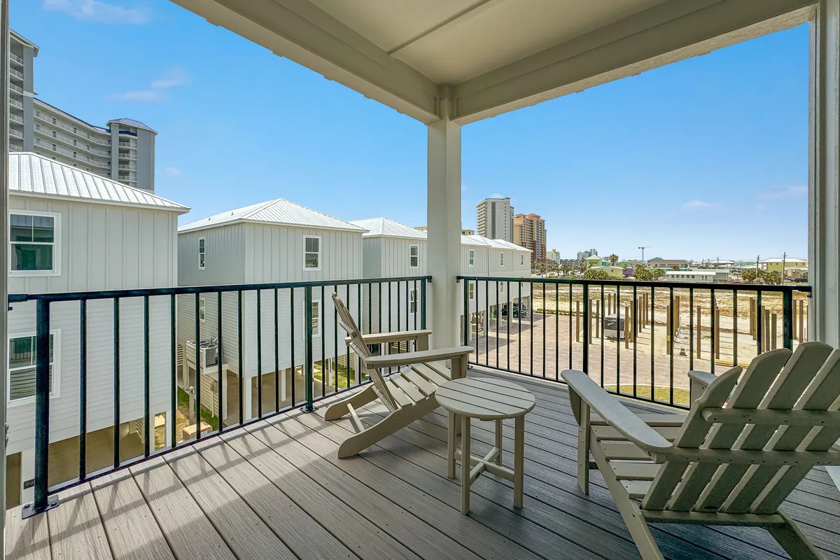 A covered balcony seating area with Adirondack chairs provides a restful vantage point overlooking neighboring homes and the urban skyline in the distance. The scene is bathed in natural light and offers a sense of outdoor relaxation and connection to the surrounding Gulf Shores neighborhood.