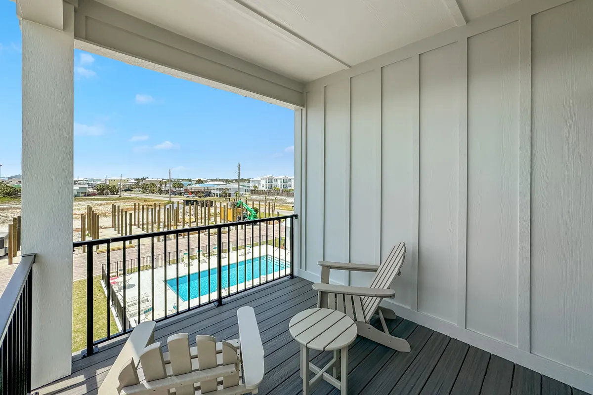 The image features a covered balcony with Adirondack chairs, offering a sweeping view of the community pool below and glimpses of the neighborhood and distant beach area. The tranquil setting encourages relaxation after a day outdoors.