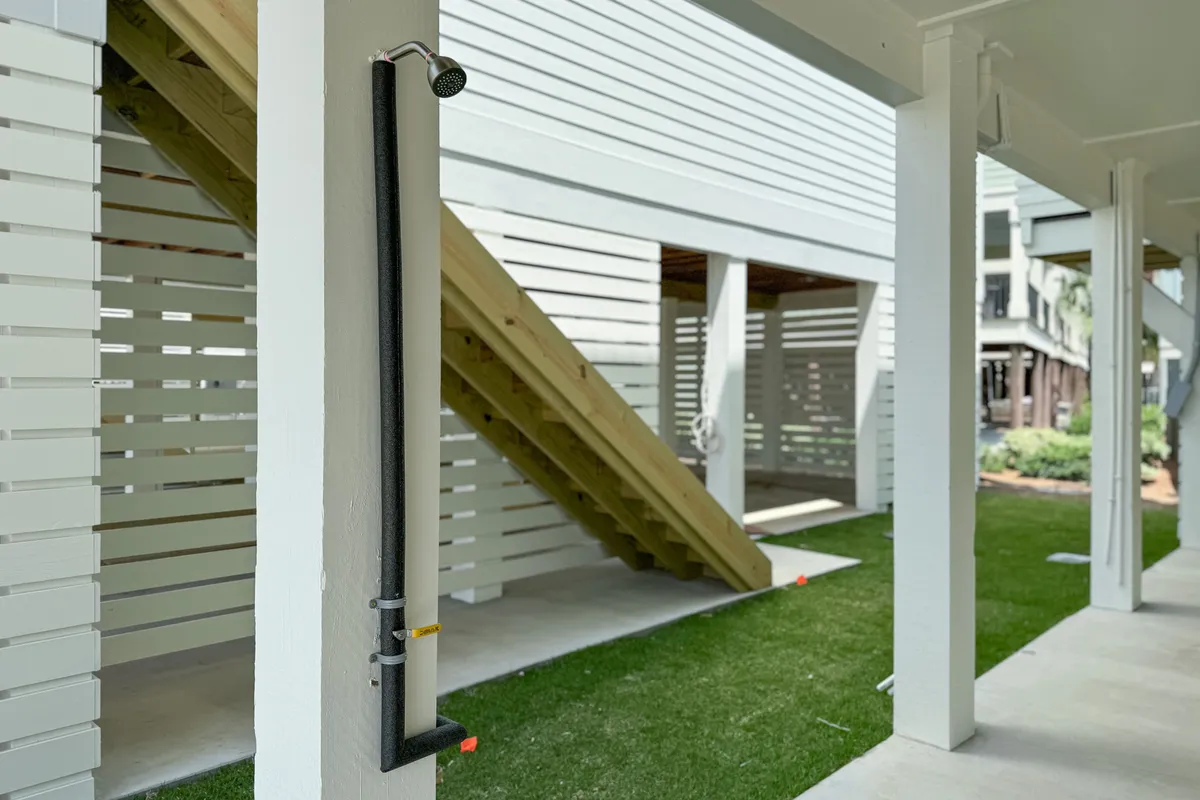 An outdoor shower is attached to a white pillar, set beside a wooden staircase and enclosed by white slatted privacy walls, with well-maintained green grass beneath the elevated home. This practical and refreshing feature makes it easy to rinse off after beach outings before heading indoors.